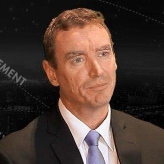 A man in a suit and tie with a serious expression, against a dark background.