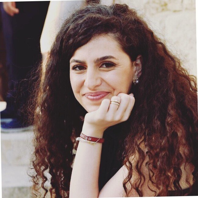 Young woman with long curly dark hair smiling, resting her chin on her hand, wearing earrings and a red bracelet.