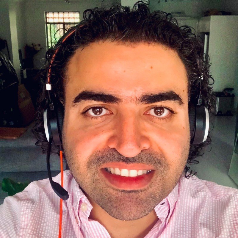 A man with curly dark hair wearing a pink checkered shirt and a headset, smiling at the camera indoors.