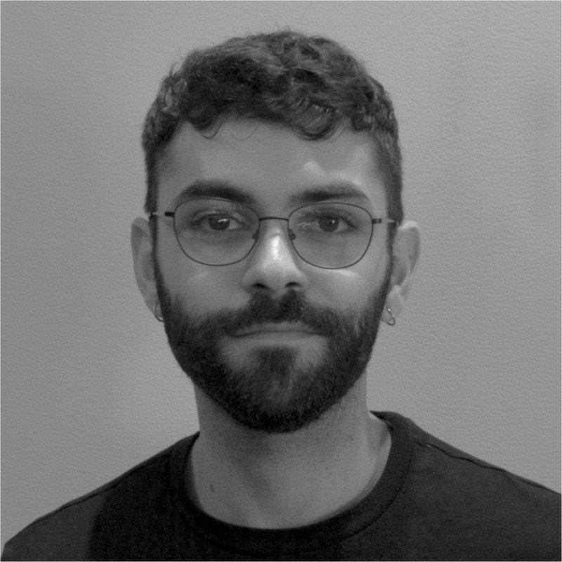 Black and white portrait of a young man with curly hair, glasses, a beard, and a small earring in his left ear, wearing a dark shirt against a plain wall background.