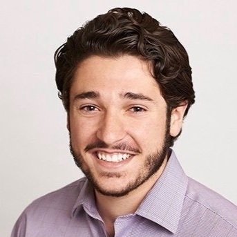 A smiling man with dark wavy hair and a beard, wearing a light purple shirt, posing against a plain background.