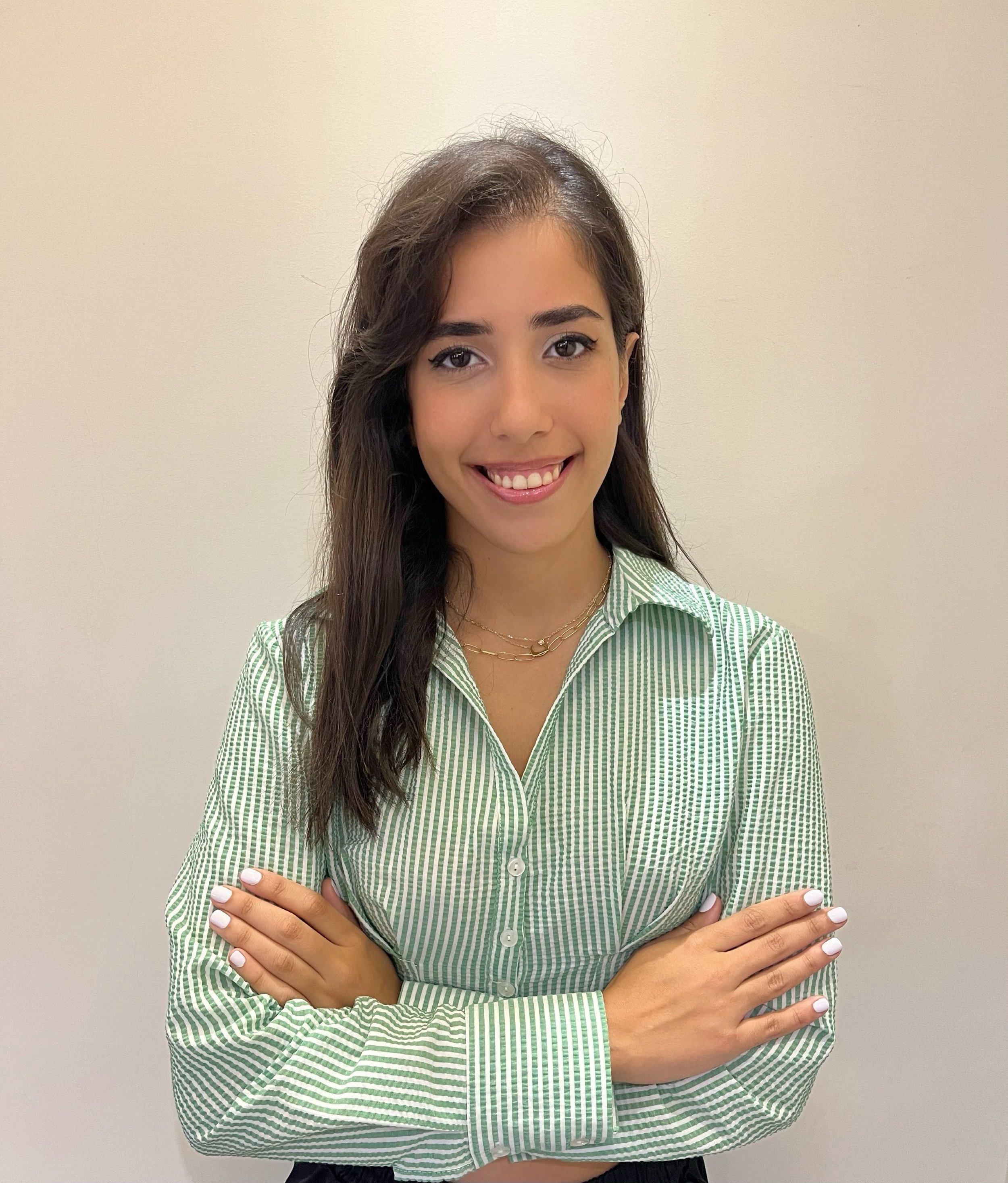 A young woman with long dark hair smiling with crossed arms, wearing a green striped button-up shirt and gold jewelry, standing against a plain beige wall.