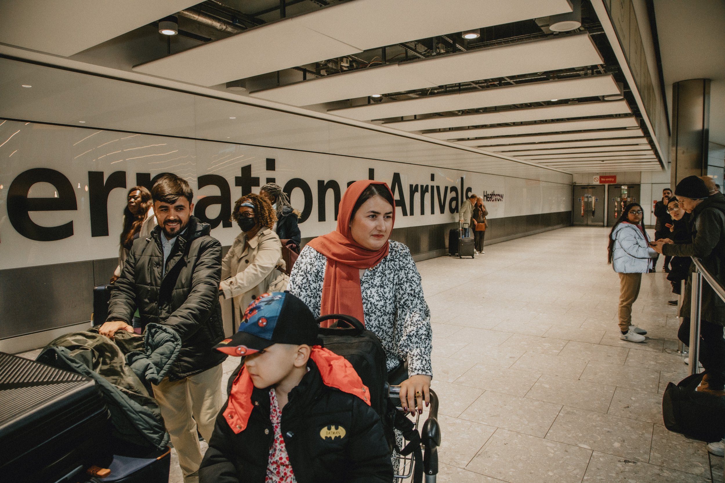 People standing and waiting in the airport arrivals area, with a large sign reading 'International Arrivals' on the wall behind them.
