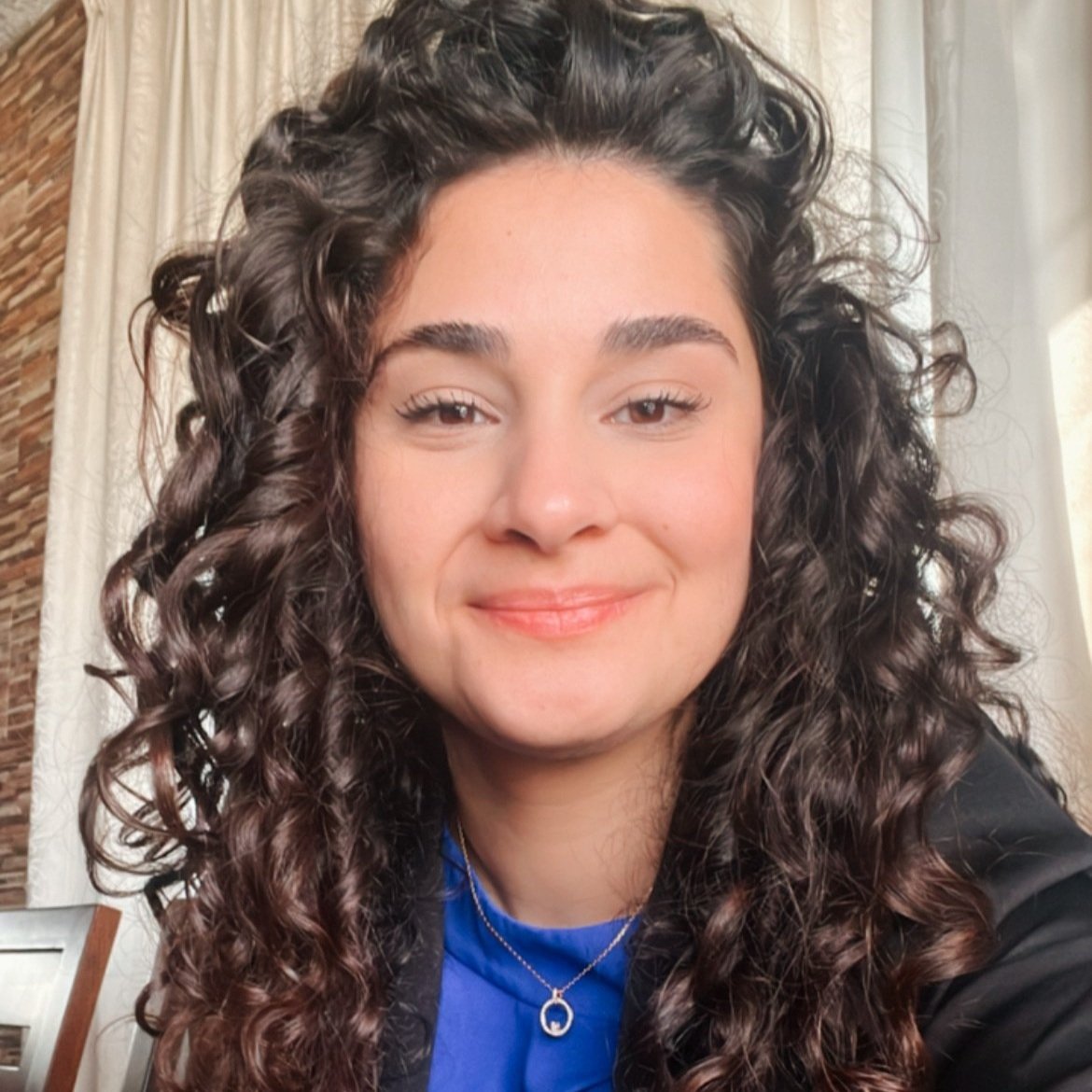 Close-up of a woman with dark, curly hair, smiling softly, wearing a blue top and a necklace, indoors with cream curtains and a brick wall in the background.