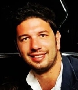 A smiling man with dark hair and a beard, wearing a white shirt and dark jacket, against a dark background.