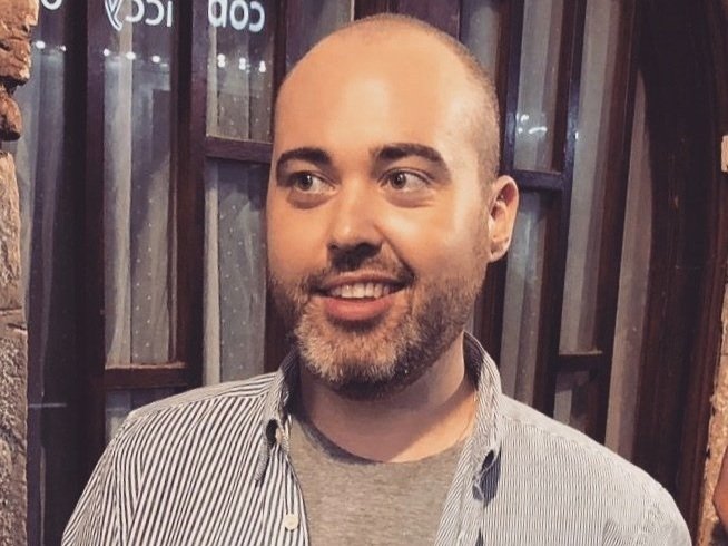 A man with a beard and a shaved head, wearing a gray t-shirt and a striped shirt, smiling and looking to the side in a setting with wooden and metal surroundings.