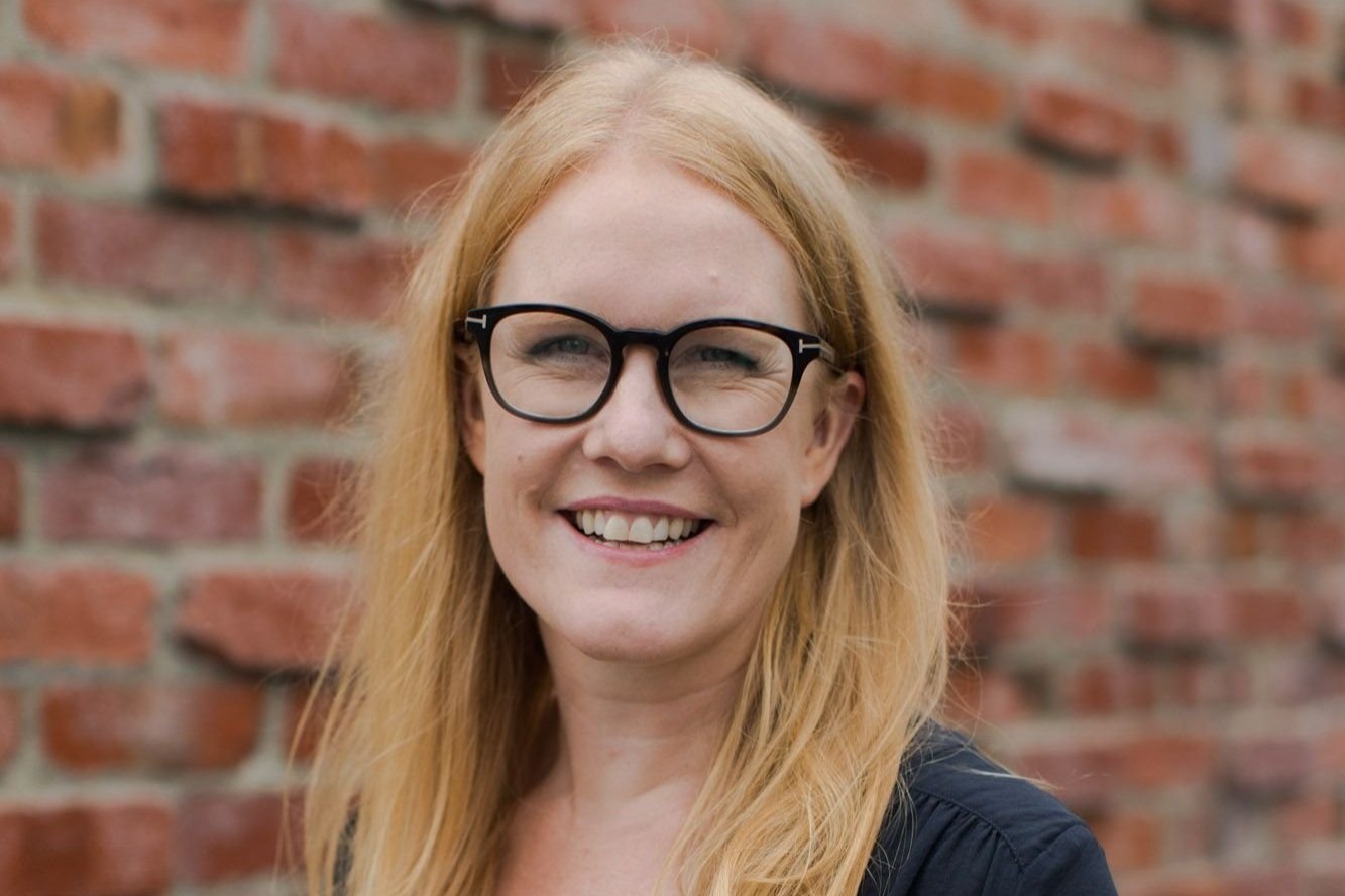 A woman with long red hair, glasses, smiling, standing outdoors in front of a red brick wall.