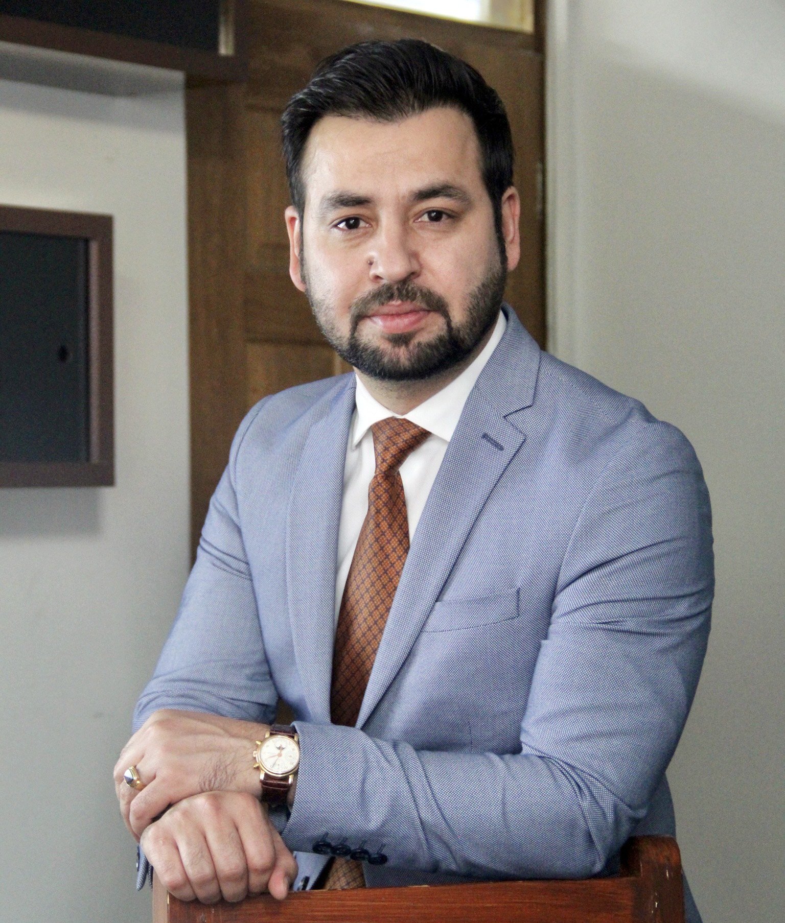 A man with dark hair, beard, and mustache wearing a light gray suit, white shirt, and brown patterned tie sits with arms crossed, looking at the camera, in an office setting.