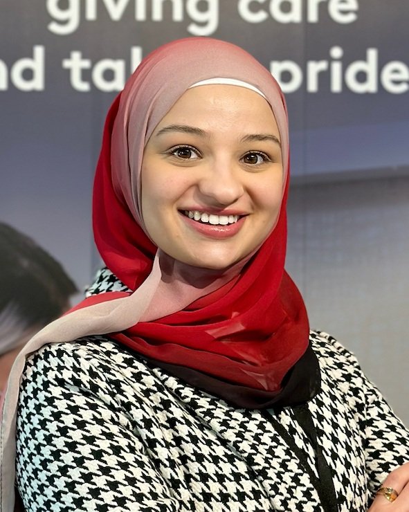 A smiling woman wearing a pink, beige, and red hijab and a black-and-white houndstooth patterned blazer.