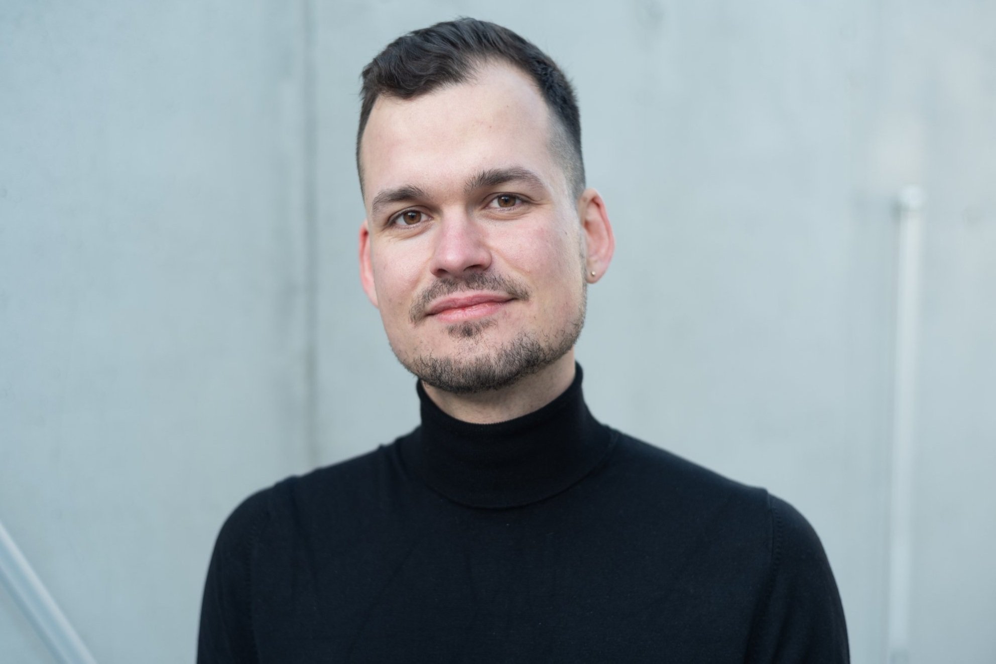 A young man with short dark hair and a trimmed beard, wearing a black turtleneck, standing in front of a plain gray wall.