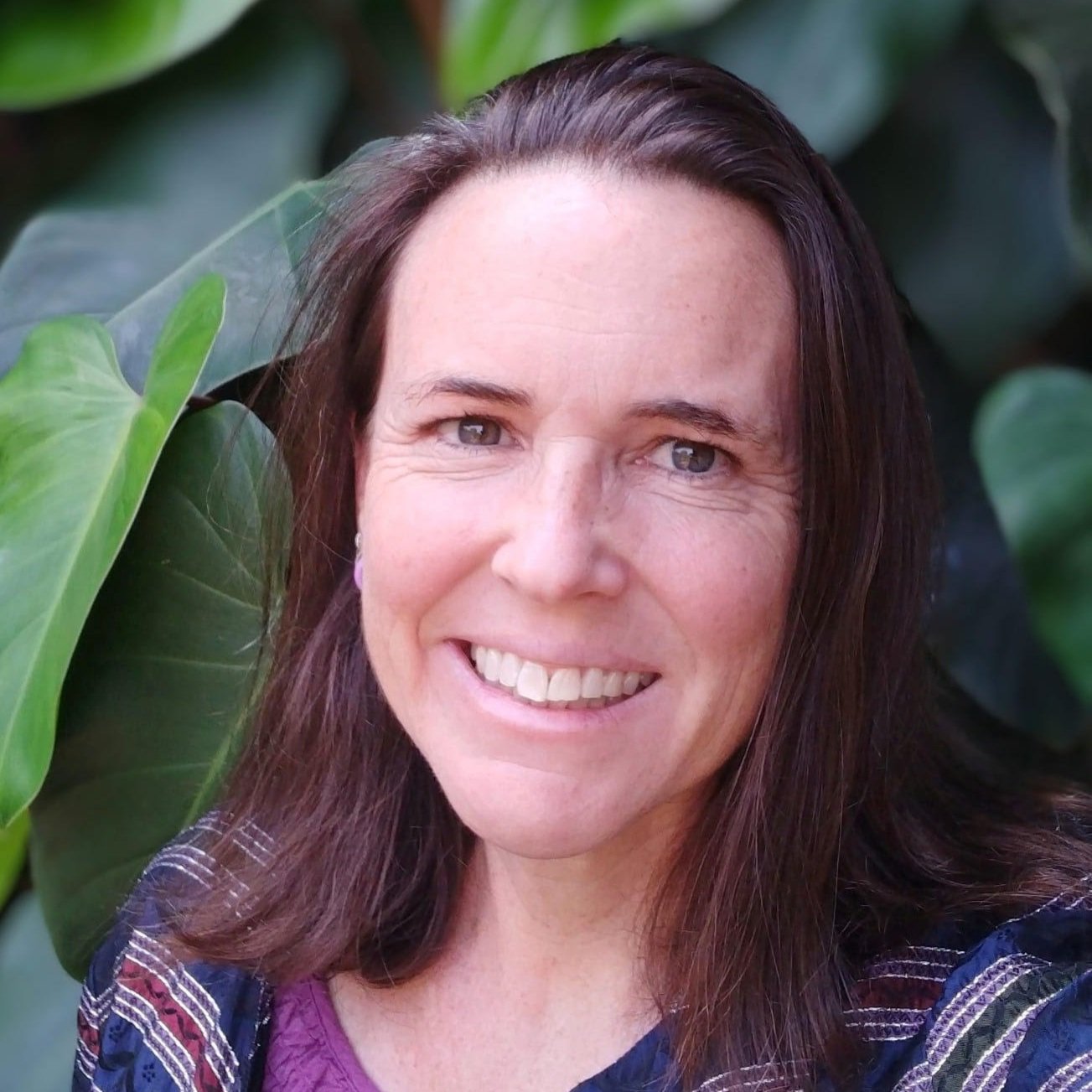 A woman with long brown hair smiling in front of large green leaves.
