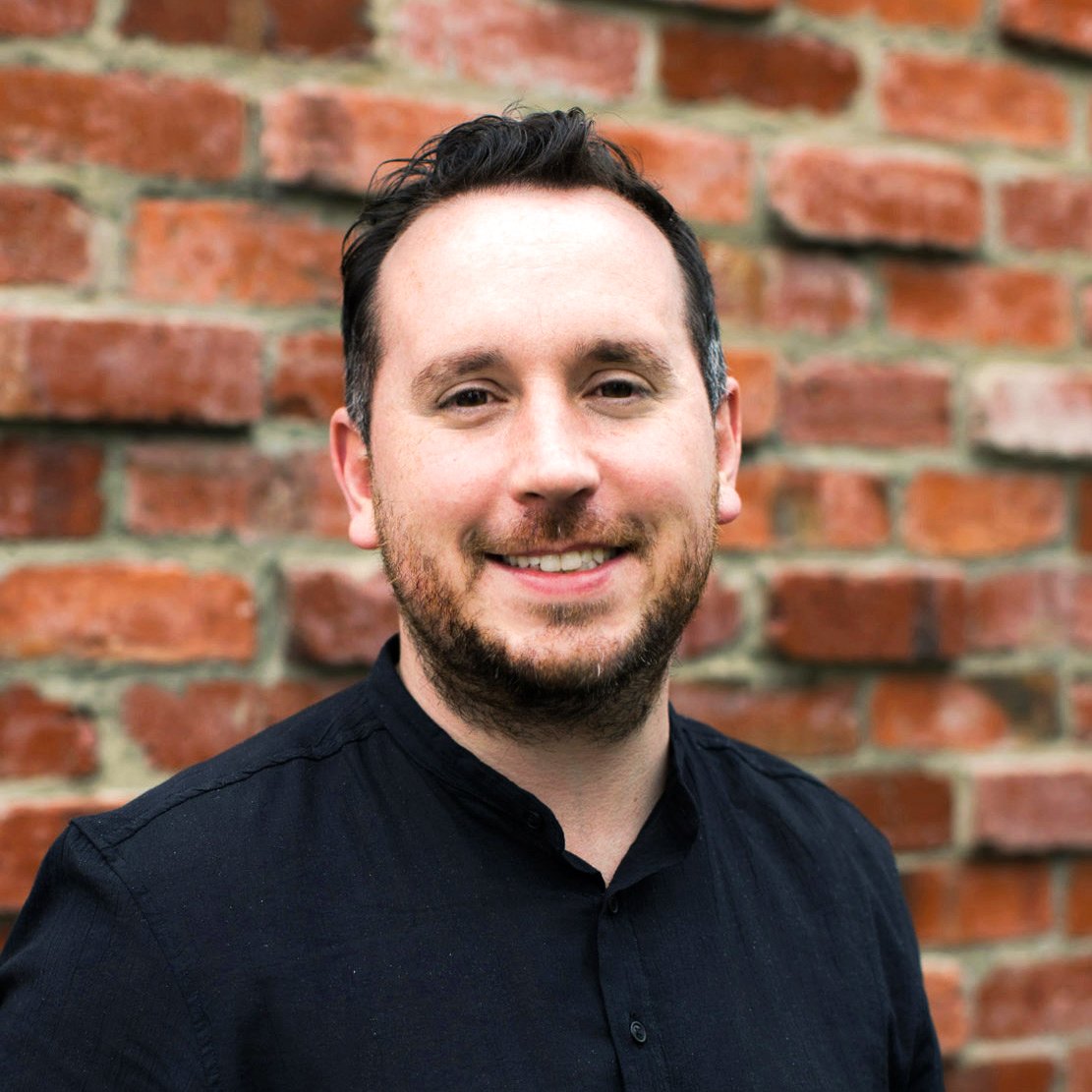 A smiling man with short dark hair and a beard, wearing a black shirt, standing in front of a brick wall.