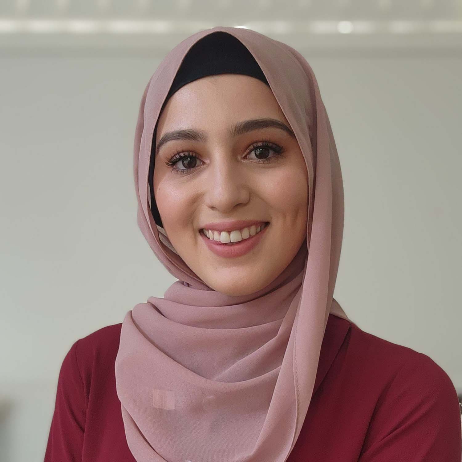 A young woman wearing a pink hijab and maroon top, smiling at the camera against a plain background.