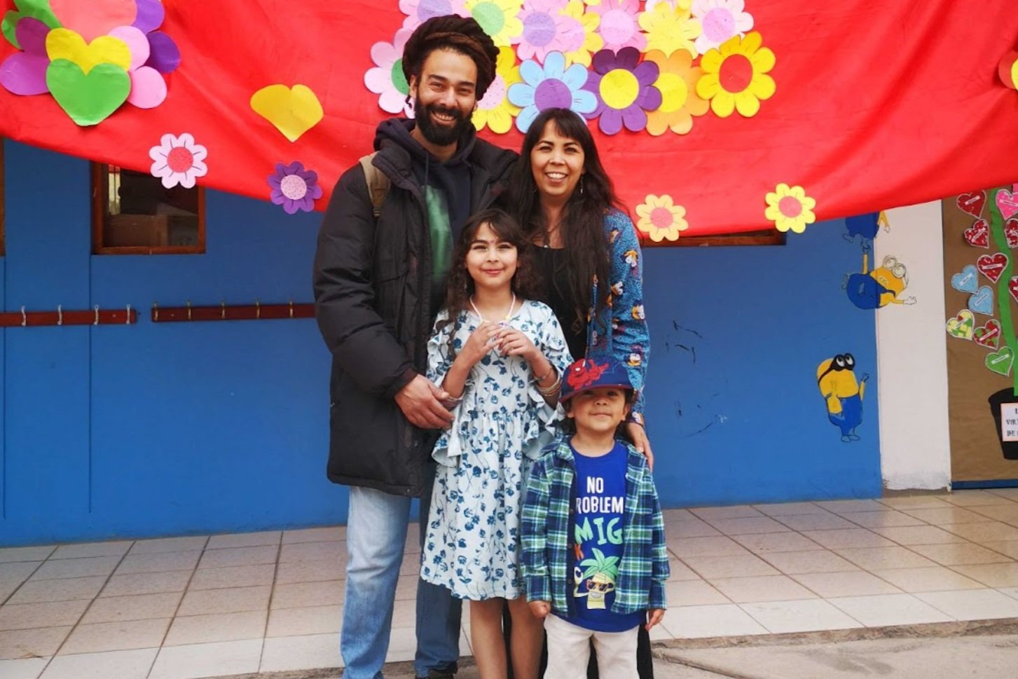 A family of four, two adults and two children, standing in front of colorful paper flowers and hearts on a red backdrop. They are smiling and posing for the photo.
