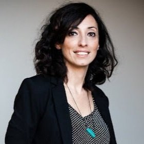 A woman with curly dark hair wearing a black blazer and top, smiling against a neutral background.