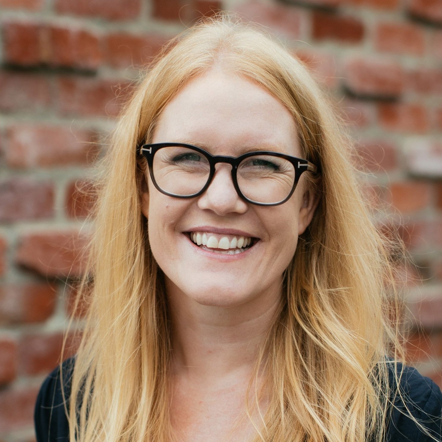 Close-up of a smiling woman with long red hair, wearing black-rimmed glasses, standing in front of a brick wall.