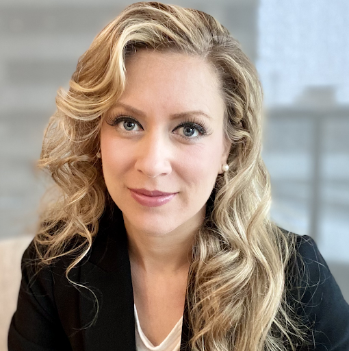 A woman with blonde, wavy hair and light skin, wearing a dark blazer and a white top, smiling slightly, in an indoor setting with blurred background.