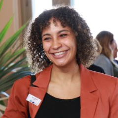 Smiling woman with curly hair wearing a burnt orange blazer and black top, standing indoors with plants and other people in the background.