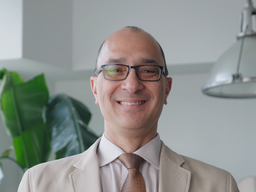 A man with glasses, balding head, and light skin, smiling at the camera, wearing a beige suit jacket, white shirt, and brown tie, indoors with a plant and a lamp in the background.